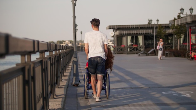 Guy rolls a disabled girl with the red hair in a wheelchair on the waterfront