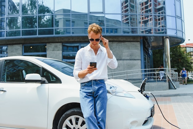 Il ragazzo si sedette sul cofano dell'auto. la sua auto è in carica alla stazione di ricarica. un uomo guarda lo schermo dello smartphone e sorride.