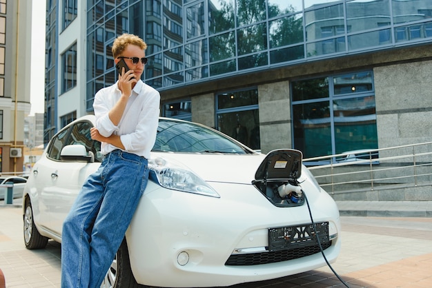 男は車のボンネットに腰を下ろした。彼の車は充電ステーションで充電しています。男はスマホの画面を見て微笑む。