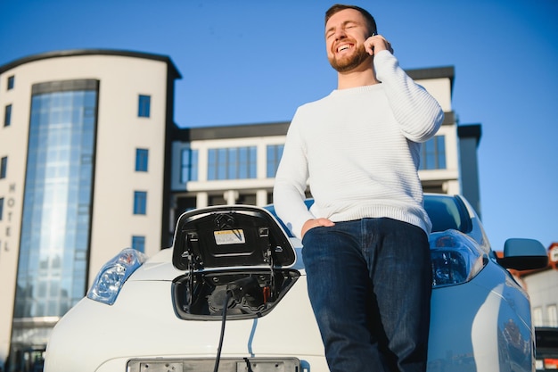 The guy sat down on the hood of the car. his car is charging at\
the charging station. a man looks at the smartphone screen and\
smiles.