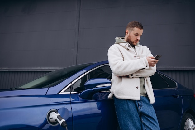 Photo the guy sat down on the hood of the car his car is charging at the charging station a man looks at the smartphone screen and smiles