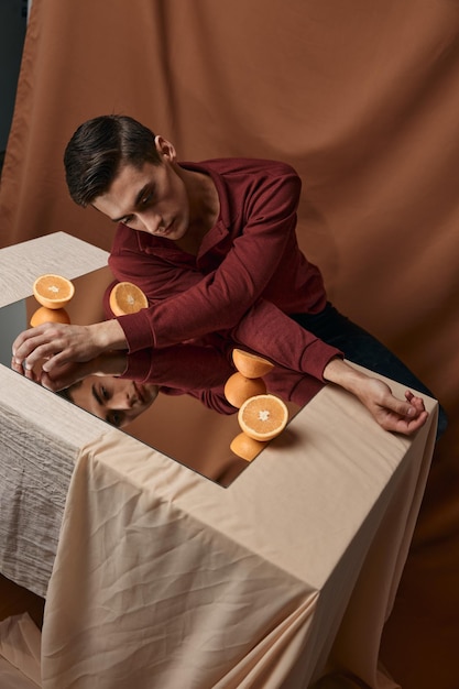 A guy in a shirt sits at a table with a mirror and orange oranges fabric background
