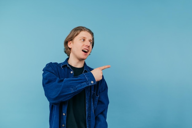 guy in a shirt stands on blue background with a smile on face looking away and pointing index finger