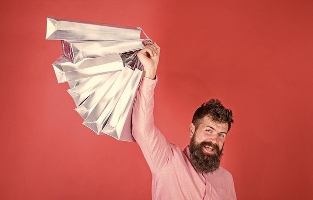 Guy shopping on sales season with discounts man with beard and\
mustache holds shopping bags red background hipster on happy face\
is shopping addicted or shopaholic shopping concept