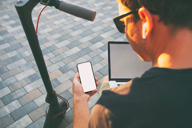 Il ragazzo seduto in panchina con scooter elettrico e laptop e che mostra il telefono con uno schermo bianco vuoto