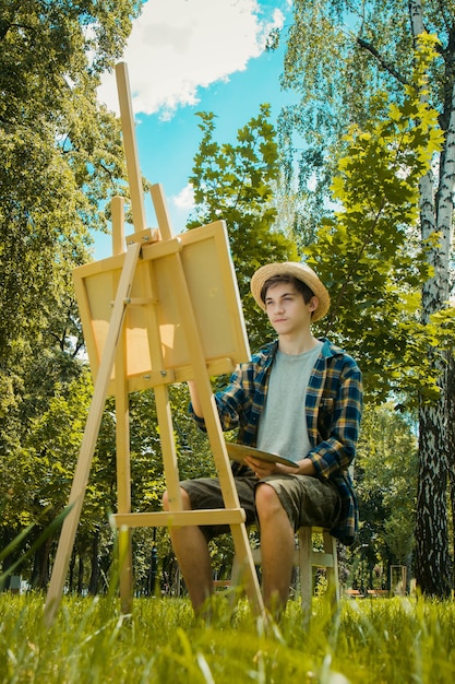 Photo guy in a straw hat sits in the park in front of an easel