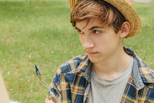 Foto ragazzo con un cappello di paglia si siede nel parco davanti a un cavalletto