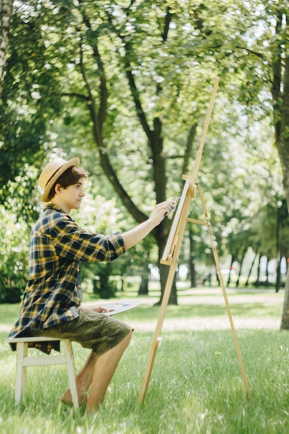 guy in a straw hat sits in the park in front of an easel