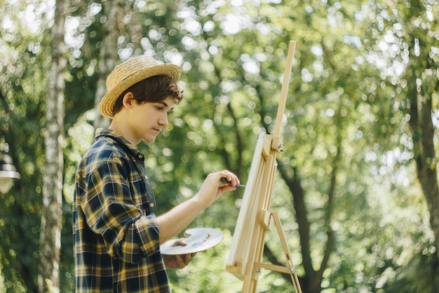 guy in a straw hat sits in the park in front of an easel