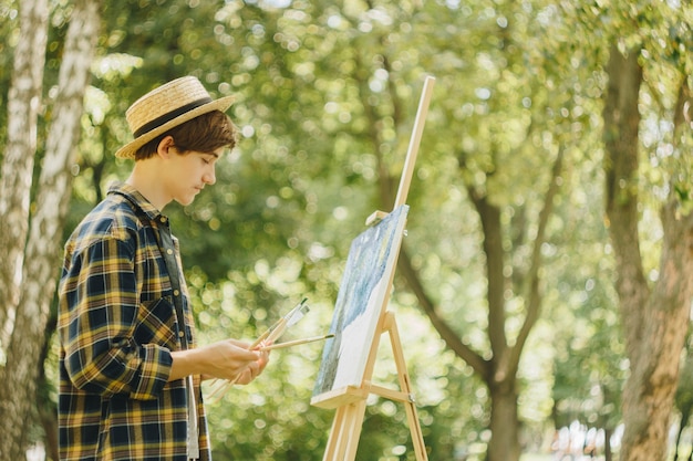 guy in a straw hat sits in the park in front of an easel