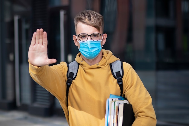 Guy student, pupil boy, young man in protective medical mask and glasses on face outdoors university with books, textbooks show palm, hand, stop no sign. Virus, pandemic coronavirus concept. Covid-19