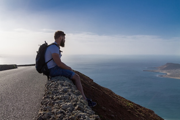 A guy in summer clothes is sitting on a stone fence and looks at a nearby island in the ocean. Mirador del Rio, Lanzarote, Spain.