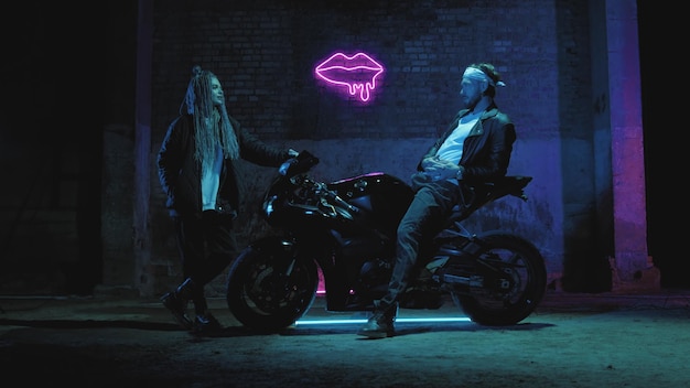 A guy on a super sport motorcycle stands talking to a girl against a pink neon sign