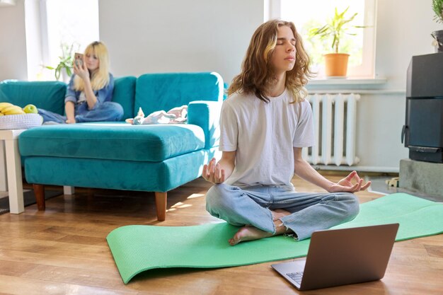 Guy tiener zittend in lotuspositie op yoga mat thuis op de vloer met laptop