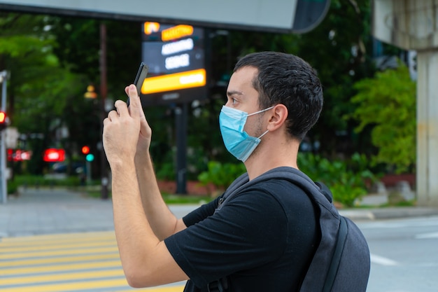 Парень турист с рюкзаком на спине и хирургической маской на лице, фотографирует здания на городской улице. Коронавирус пандемия. Последствия для туризма.