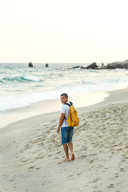 黄色のバックパックを持つ男の観光客は、海沿いの砂浜に沿って歩きます。