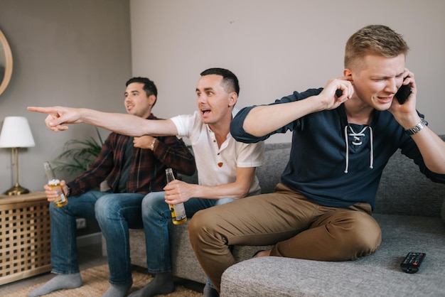 Guy trying to talk on the phone, but his friends distracting him from the phone. Group of friends watching a football match on TV against the background of young man phone conversation.