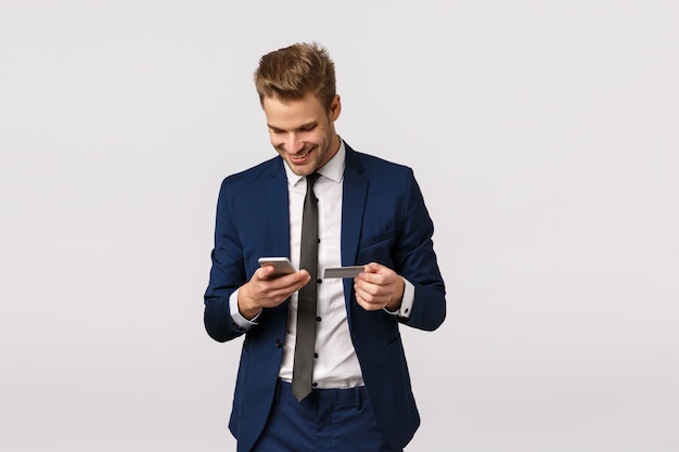 Guy want buy flowers for girlfriend, working office, holding smartphone and credit card, smiling ad enter billing details display, standing white background. Businessman paying for purchase