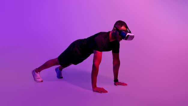 Guy wearing vr glasses doing plank exercise over purple background