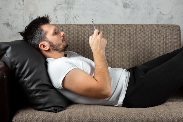 guy wearing a white shirt is lying on the couch and sitting in the phone. 