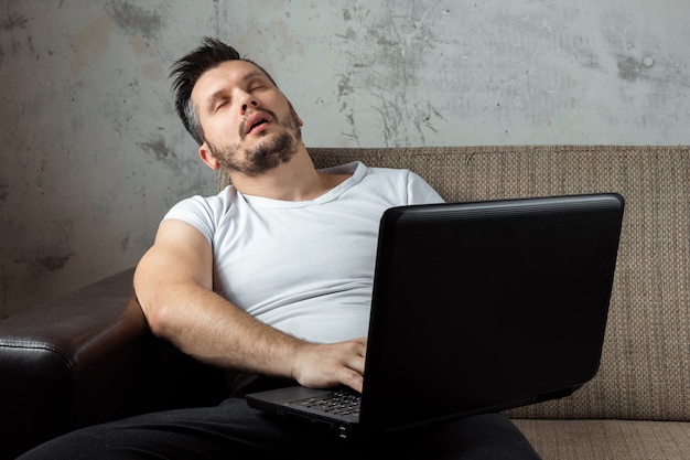 guy wearing a white shirt sitting on the couch, fell asleep at work on a laptop. 