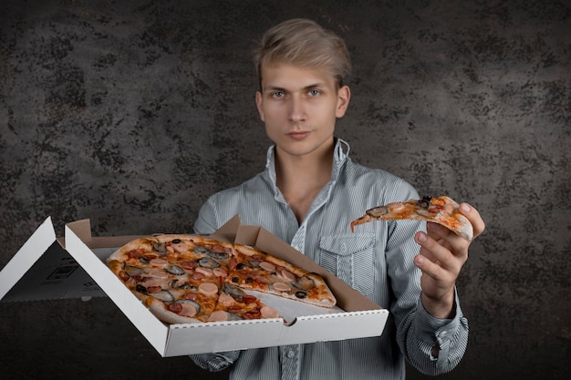 Un ragazzo con una maglietta bianca tiene in mano un pezzo di pizza su uno sfondo nero. giovane che mangia una fetta di pizza con gli occhi chiusi, isolato su sfondo giallo.