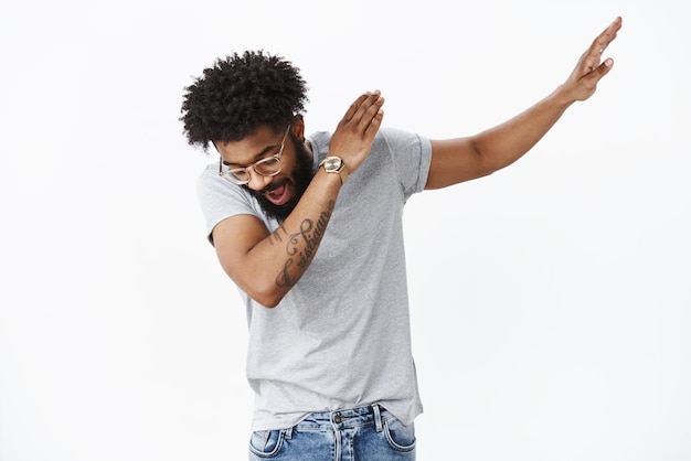 guy with beard and tattoos sweep hand right and looking down as making dance move, shaking and having fun on gray wall.