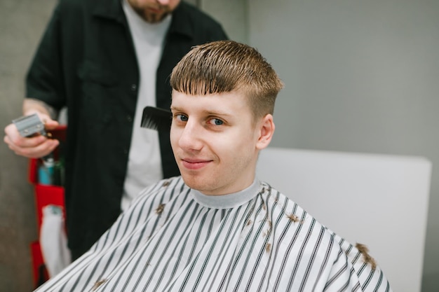 Guy with blond hair cuts his hair at a professional barber looks in camera with a smile on his face