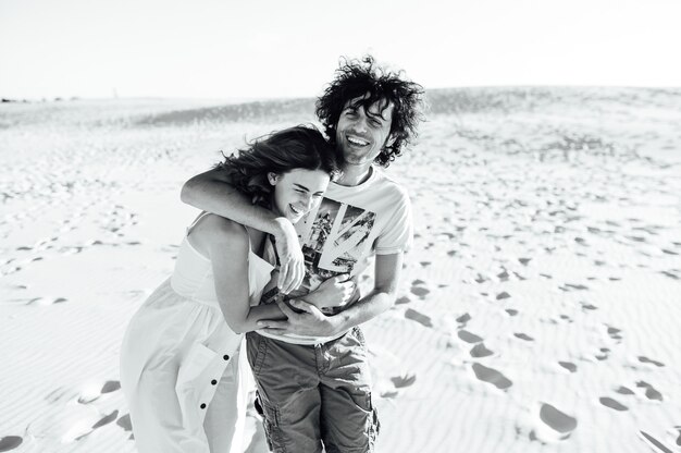 Un ragazzo con i capelli ricci e la sua ragazza si stanno abbracciando allegramente sulla sabbia.