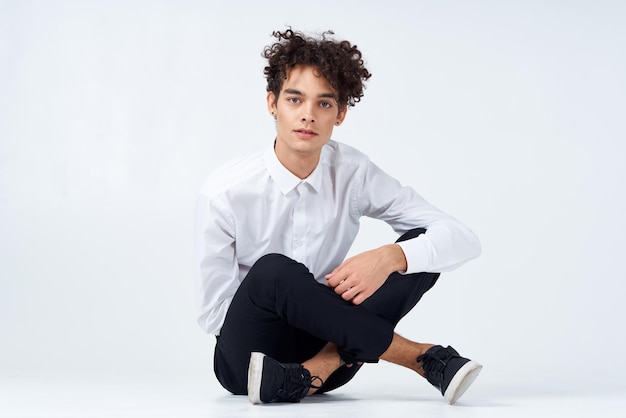 A guy with curly hair in a white shirt sneakers and trousers sits on the floor In a bright room