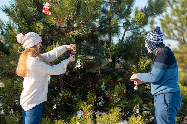Парень с девушкой украшают зеленую елку на улице зимой в лесу декоративными игрушками и гирляндами, елочными игрушками