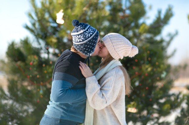 冬の森の中でお祝いおもちゃや花輪で飾られた緑のクリスマスツリーの女の子のキスを持つ男。冬のロマンス