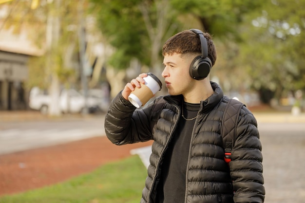 コピースペースのある公共の公園を歩きながら、ヘッドフォンをした男性がコーヒーを飲む