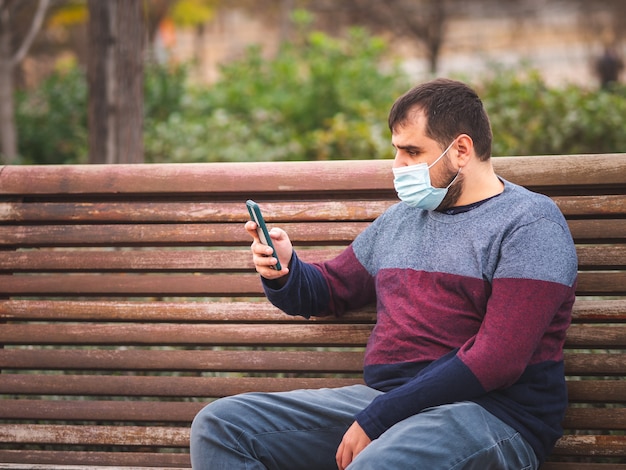 日没時に公園のベンチで彼のスマートフォンを使用して保護マスクを持つ男