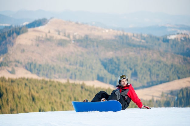 Фото Парень со сноубордом сидит на снежном склоне на вершине горы и смотрит в сторону