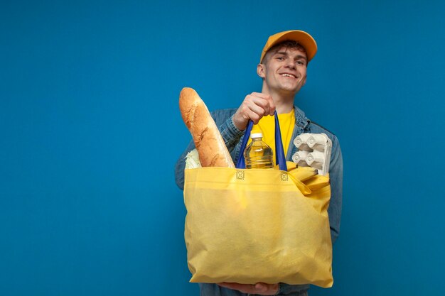 Guy in yellow cap holds fabric eco bag full of food and smiles on blue background