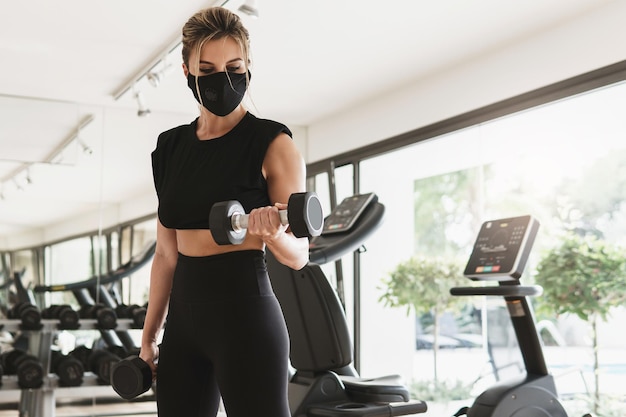 Palestra nuova normalità. giovane donna atletica che indossa una maschera facciale di prevenzione durante il suo allenamento fitness con manubri.