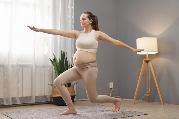 Фото Гимнастика во время беременности спортивная беременная женщина в бежевом топе и леггинсах в светлом интерьере гостиной практикует йогу, наслаждается домашней спортивной деятельностью