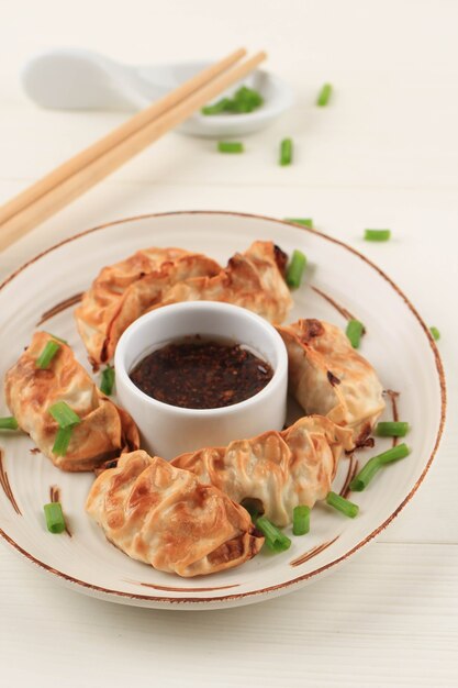 Gyoza азиатские Potstickers с соусами, подаваемые в коричневой керамической тарелке в японском стиле с палочками для еды и весенним луком на белом деревянном фоне. Приготовление пищи в аэрогриле без масла.