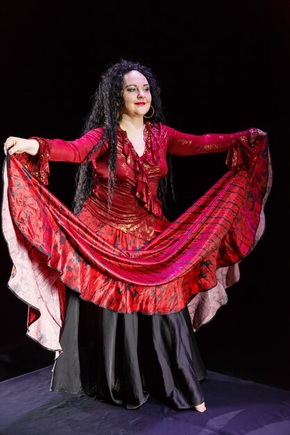 Gypsy woman barefoot with long black hair dances waving\
red-black skirt on a black surface