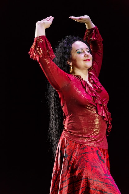 Gypsy woman with long black hair dances with arms with sleeves\
in a red dress on a black background.