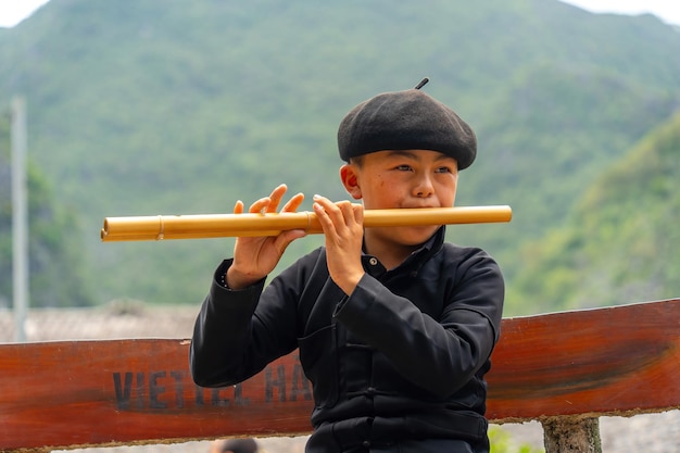 Ha Giang VIETNAM 10 augustus 2023 Portret van een schattige kleine jongen die een zwart t-shirt draagt en fluit speelt Muzikant jonge jongen die zholomiya etnisch houtblazers muziekinstrument speelt