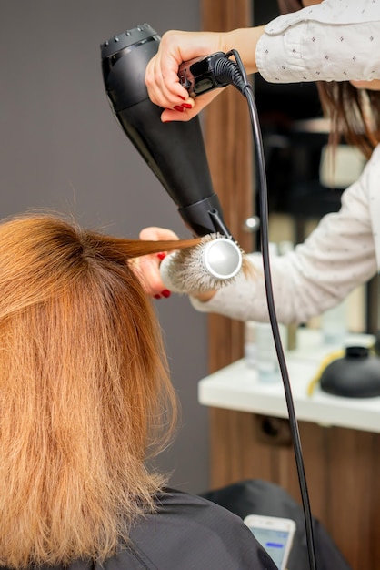 Haar drogen in de haarstudio. Vrouwelijke kapper stylist droogt haar met een haardroger en ronde borstel rood haar van een vrouw in een schoonheidssalon.