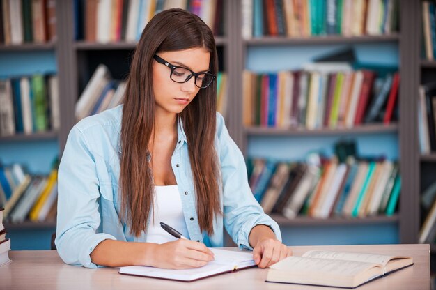 Haar huiswerk maken. Jonge vrouw schrijft iets in haar notitieblok en leest een boek terwijl ze aan het bureau in de bibliotheek zit