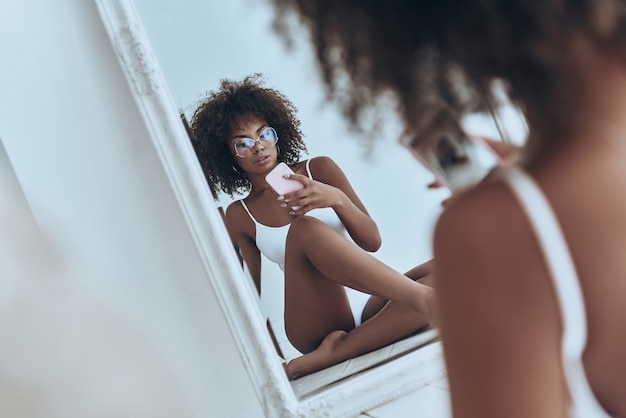 Haar lichaam is perfectie. Weerspiegeling van mooie jonge Afrikaanse vrouw in witte lingerie die selfie in de spiegel neemt terwijl ze thuis op de vloer zit