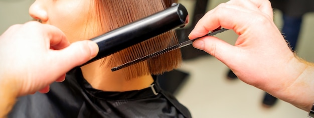 Haarstylisten handen rechttrekken kort haar van jonge brunette vrouw met stijltang en kam in een be...