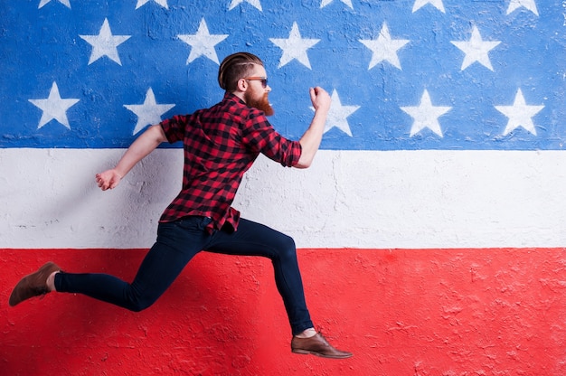 Haast om in de trend te zijn. Knappe jonge bebaarde man die een zonnebril draagt en langs de Amerikaanse vlag loopt