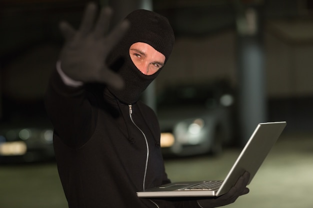 Hacker in balaclava gesturing and using laptop