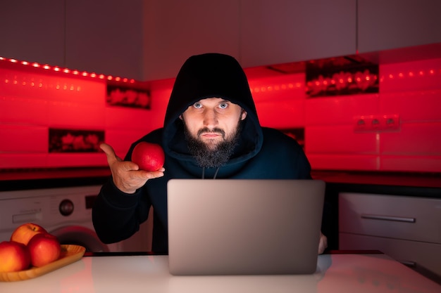 A hacker in a black hoodie with a hood on his head is sitting\
at a laptop holding a red apple in front of him