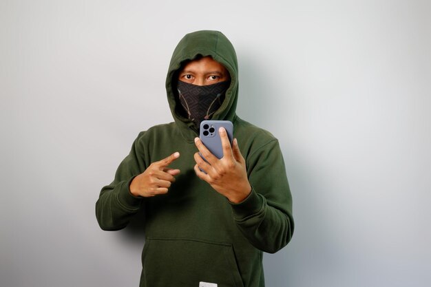 Hacker die naar de camera kijkt, buff en groene hoodie draagt, een mobiele telefoon vasthoudt en er naar wijst